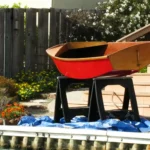 A small wooden boat with red boat paint on its hull, sitting on sawhorses in a backyard, ready for finishing work.