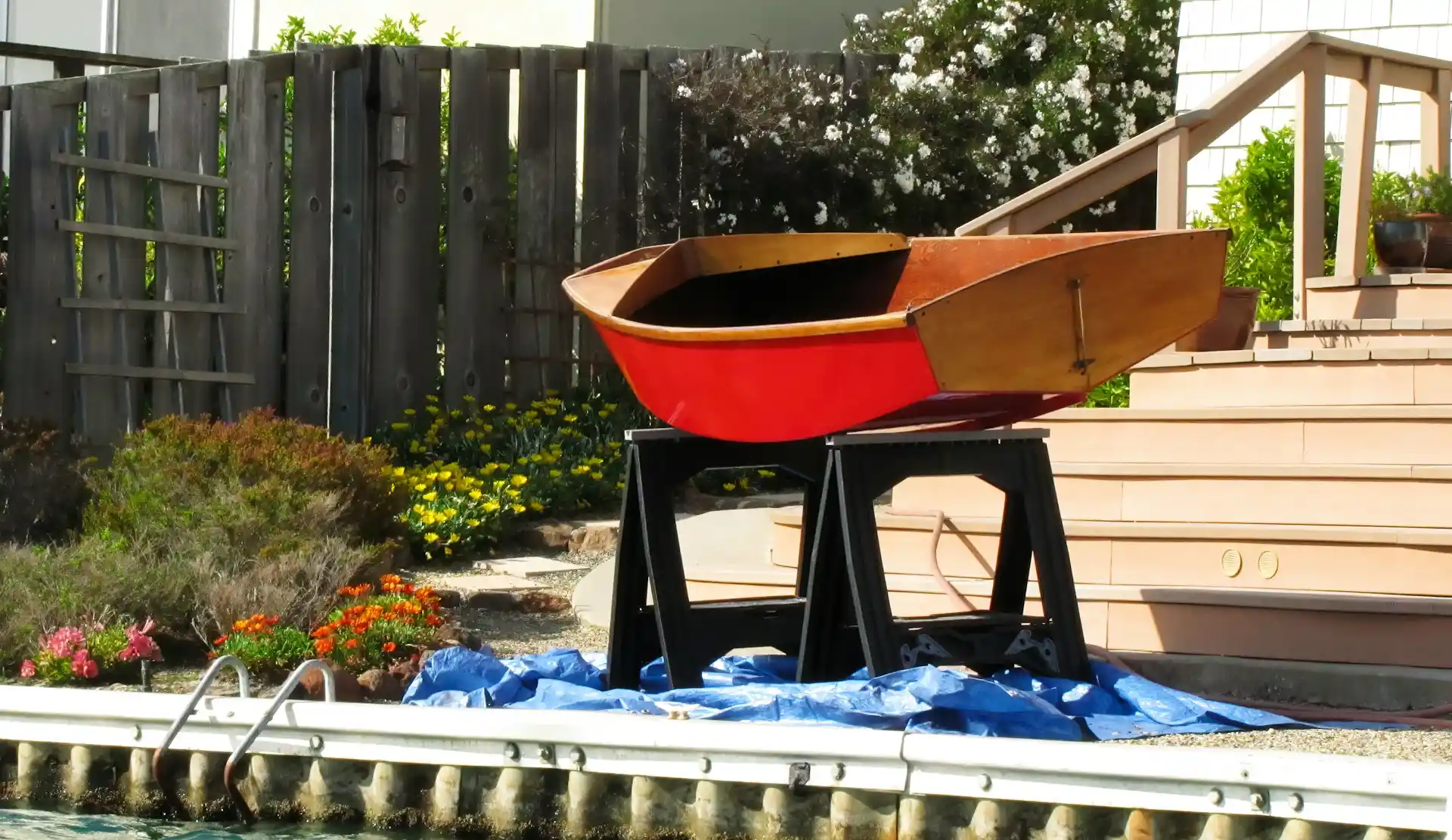 A small wooden boat with red boat paint on its hull, sitting on sawhorses in a backyard, ready for finishing work.
