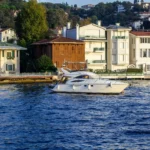 A modern white yacht moving on blue water with houses on the shore, prompting consideration of houseboats expensive costs and ownership expenses.