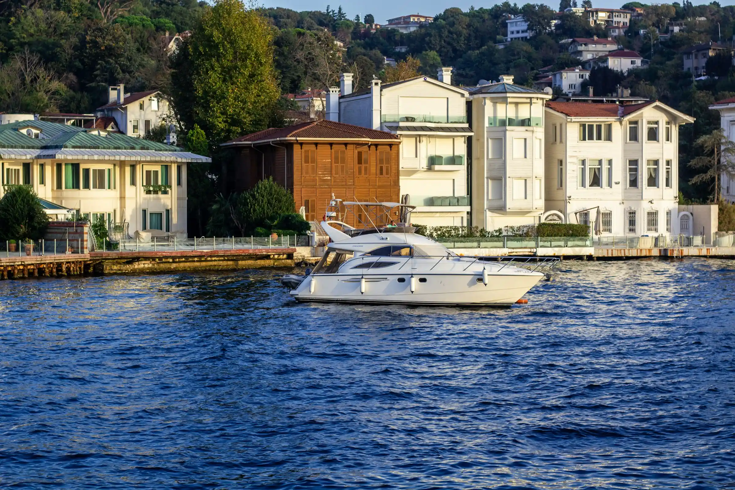 A modern white yacht moving on blue water with houses on the shore, prompting consideration of houseboats expensive costs and ownership expenses.