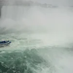 A small tour boat navigating turbulent, misty waters at the base of a powerful waterfall, symbolizing the importance of houseboat storm safety.