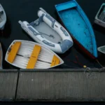 An overhead view of several small boats of various designs and colors, including wooden and inflatable, docked in dark water next to a wooden pier, illustrating a range of boat types throughout the evolution of boat paint.