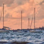 Three sailboats anchored in a calm bay at sunset, their silhouettes against a dramatic sky, prompting questions about whether do houseboats hold their value.
