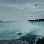 A small boat navigating the turbulent waters at the base of a massive waterfall, symbolizing the powerful forces that might lead one to ask: Can houseboats sink?