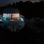A houseboat illuminated by interior lights at night, docked in dark water with a treeline in the background, visually demonstrating how do houseboats get electricity.